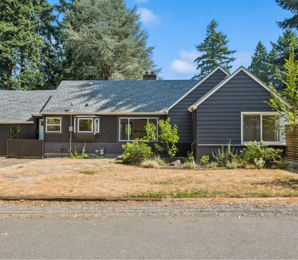 Front view of a dark-colored house with a sloped roof, surrounded by greenery and dry grass, illustrating properties available for quick cash offers from Portland Cash Buyers.
