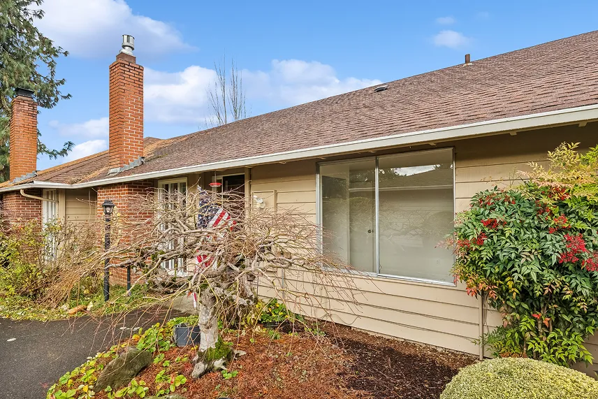 Exterior view of a single-story house with a brown roof, brick chimney, and landscaped front yard featuring shrubs and decorative plants, relevant to Portland Cash Buyers' service of purchasing homes as-is.